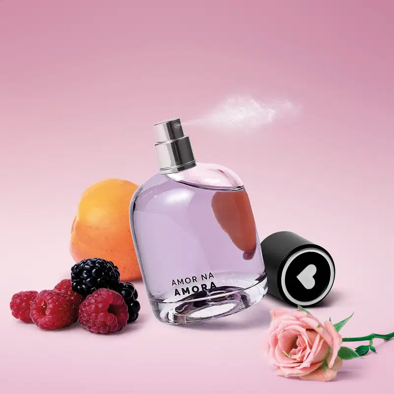 Fragrância em frasco de vidro. Ao redor, uma laranja, amoras vermelhas e pretas, uma rosa e uma tampa preta com um símbolo de coração branco. As cores predominantes são rosa, preto, vermelho e laranja. Sugere uma fragrância com notas frutadas e florais.