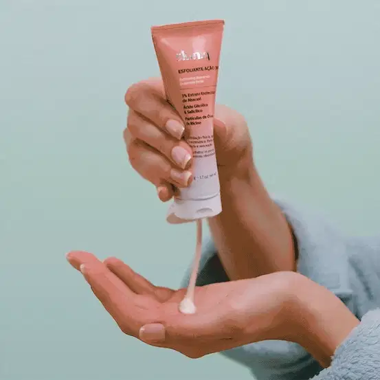 Mão segurando uma bisnaga laranja e branca com letras pretas de esfoliante facial Skin.q Ação 360° da marca Quem Disse, Berenice? e apertando o frasco, derrubando uma porção do produto na outra mão. O fundo da imagem é azul.
