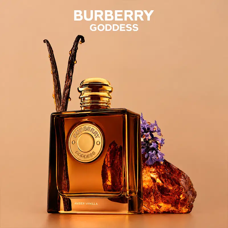 Perfume Burberry Goddess em frasco quadrado âmbar com tampa dourada. Rótulo circular dourado com inscrição "Burberry Goddess". Acompanhado por vanilla amber e elementos decorativos naturais em tom dourado e roxo.